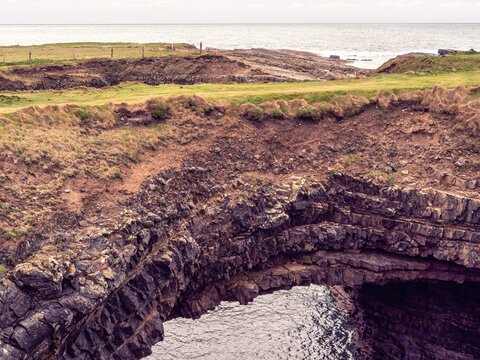 Natural rock arch over calm ocean water. Rocky cliffside with grassy edges. Weathered stone forms a curved tunnel shape. Calm sea beneath reflects sky light. Rosses Point, county Clare, Ireland.