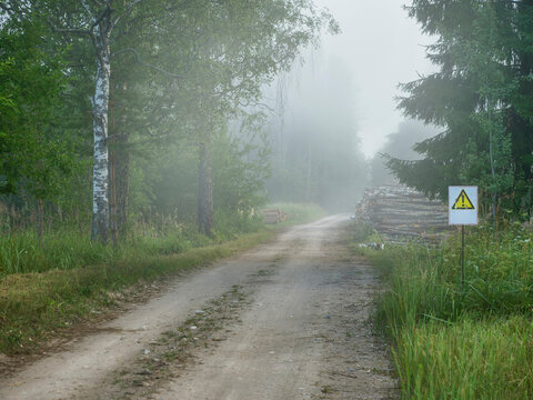 Misty forest path with construction warning sign. Gravel road winds through dense green trees. Fog partially obscures distant trees. Sign attention mark. Pile of wood on a side of the road.