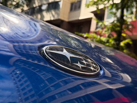Close up view of a blue Subaru car hood with its classic oval starry emblem, shiny chrome logo reflecting city buildings on glossy metallic paint, Japanese automobile brand identity outdoor portrait.