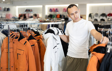 Among large selection of clothes on rails in store, guy stopped near showcase with outerwear, holds jacket hanger in hands and examines product, integrity of garniture. © JackF