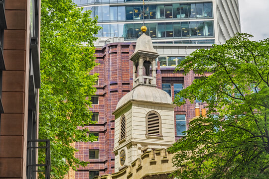 View of the historic steeple of St. Mary-le-Bow church framed by lush green trees and modern glass office buildings in London, England, United Kingdom.
