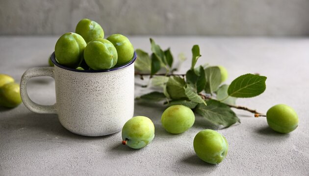 fresh green plums leafy fruit branch and speckled ceramic mug on textured grey plaster surface calm muted natural rustic scene for food blog kitchen decor and organic produce design