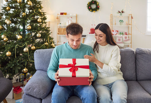Christmas Eve. Young man eagerly opens Christmas present and looks inside box with sincere admiration. Joyful woman sits next to her husband on sofa and watches with warm smile, enjoying moment.