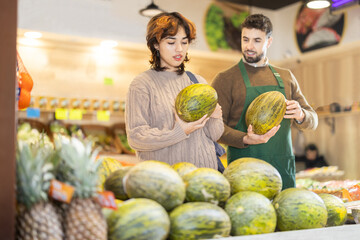 Obraz premium Girl customer in vegetable store examines melon, male seller advises and help with choice, offers ripe fruit options. Vegetable shop near house. Organic vegetable shop offer assortment