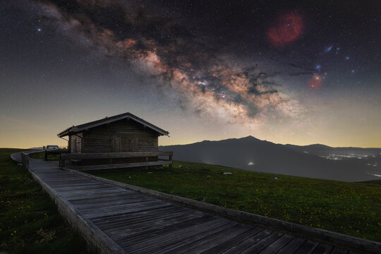 View of a wooden cabin and walkway at Forca di Presta under the starry Milky Way galaxy in the mountains of Arquata del Tronto, Marche, Italy.