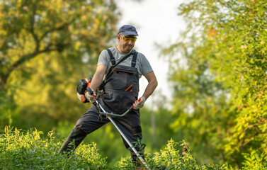 A gardener is using a weed trimmer to clear tall grass in a field. He is focused on his task, surrounded by plants and trees. The sun is shining and it is daytime © Віталій Б.