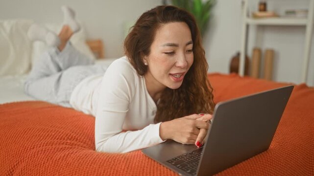 Woman lying on bed on stomach with legs raised, smiling and typing on laptop on an orange blanket in bedroom; serene comfort.
