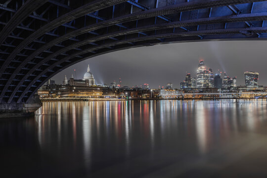 View of St Paul's Cathedral and the illuminated London skyline reflected in the River Thames at night, framed by the dark steel structure of a bridge in London, England, United Kingdom.