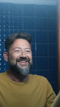 Handsome mature man looking at himself in the mirror and smiling, feeling confident and happy. Morning routine focused on self-care, self-love and building self-esteem in his bathroom