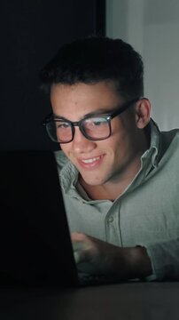 Concentrated young man wearing eyeglasses typing on a laptop computer in a dark room, with the screen light illuminating his face as he smiles, showcasing a dedicated professional working late