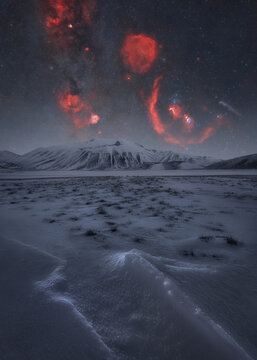 View of the snow-covered landscape of Castelluccio under a starry night sky with vibrant red nebulae above the mountains in Castelluccio, Umbria, Italy.