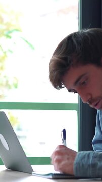 Focused man concentrating while working on his laptop, pausing to jot down notes in a notebook before resuming typing, multitasking at his desk in a modern office with a window view