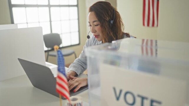 Woman holds headset mic and types on laptop beside a vote box and american flag in a building; civic pride.