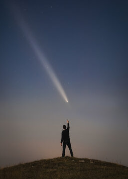 View of a person standing on a hill at night pointing at a bright comet in the starry sky above the landscape of Castelluccio, Umbria, Italy.