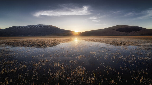 View of the sunrise over the plains of Castelluccio with reflective water and mountains in the background in Castelluccio, Umbria, Italy.