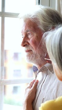 Pensive senior man looking out the window while his supportive wife joins him, offering a comforting hand on his shoulder in a tender moment of togetherness and care during a difficult time