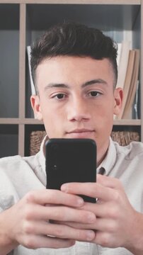 Handsome young student concentrating on his smartphone, scrolling and texting on social media or playing an online game in front of a modern bookcase, enjoying digital communication