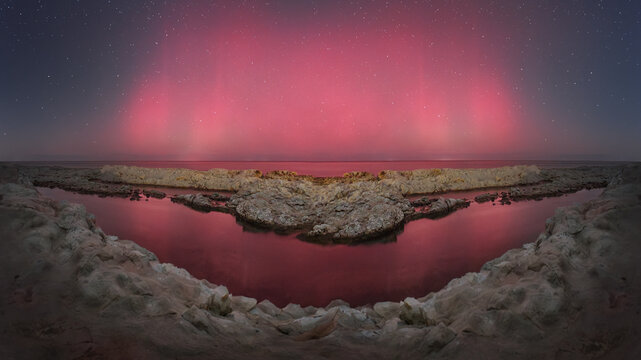 View of the Sedia del Papa rock formation on the coast of Ancona illuminated by a vibrant red aurora borealis reflecting on the calm sea water at night, Ancona, Marche, Italy.
