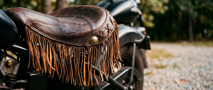 Close-up of a custom motorcycle leather seat with fringe detailing. Vintage cruiser design and biker lifestyle concept with outdoor road setting and copy space.