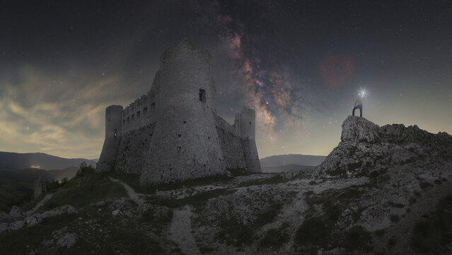 View of Rocca Calascio fortress illuminated under the Milky Way galaxy with a person standing on a nearby rock with a headlamp in Calascio, Abruzzo, Italy.