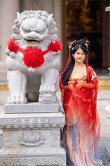 Obraz premium An elegant young woman in an ornate red hanfu costume, stands beside stone lion, hand on hip, at stone guardian lion statue with red pompom, marking the Lunar New Year in festive red attire.