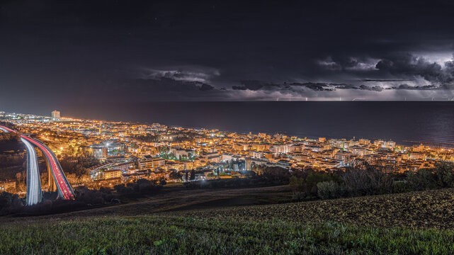 View of the coastal town of Porto San Giorgio at night, featuring illuminated highway light trails, stormy clouds over the Adriatic Sea, and city lights, Porto San Giorgio, Marche, Italy.