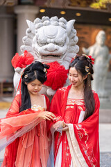 Obraz premium Wishing luck and prosperity for the Lunar New Year: two elegant young women in traditional red Chinese hanfu, hold hands, look at each other smiling, beside stone guardian lion statue with.