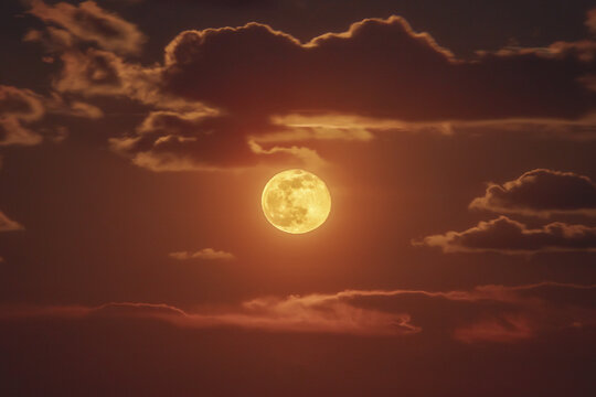 View of a bright full moon shining through dark, dramatic clouds in the evening sky in Italy.