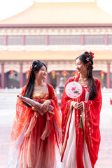 Obraz premium Wishing luck and prosperity for the Lunar New Year: two elegant young women in matching red hanfu costumes, one leaning into partner's shoulder laughing, beside temple courtyard plaza.