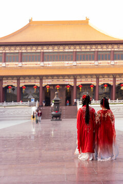 Two elegant young women wearing embroidered red hanfu robes, tighter crop, heads turned toward each other, at temple courtyard, columned facade, in a traditional hanfu culture photo shoot.