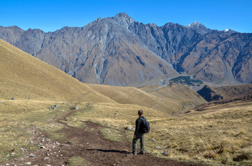 Fototapeta premium Male hiker with a backpack walks a rugged trail through a vast alpine valley in Georgia, facing massive Caucasus mountain peaks and a distant Gergeti Trinity Church under a clear blue sky