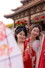 Obraz premium Portrait of two young Chinese women in red silk hanfu dresses: lowers parasol, tilts heads together smiling, framed by temple eave with red lanterns, celebrating Chinese New Year traditions.