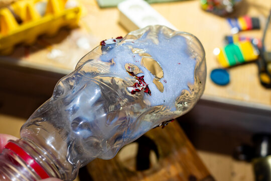 Deformed transparent plastic bottle melting under intense heat from a thermal gun, creating abstract warped textures and glossy surface patterns in a craft workshop setting