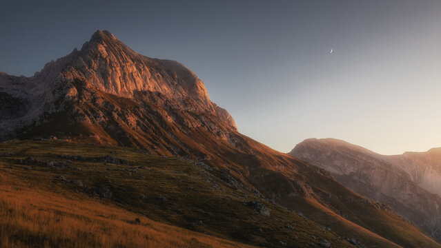 View of the Gran Sasso massif at Prati di Tivo during golden hour, with rugged mountain peaks, grassy slopes, and a crescent moon in the clear sky, Prati di Tivo, Abruzzo, Italy.
