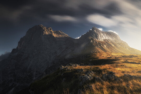 View of the dramatic rocky peaks of the Gran Sasso massif at sunset with golden grassy slopes and moody clouds in Prati di Tivo, Abruzzo, Italy.