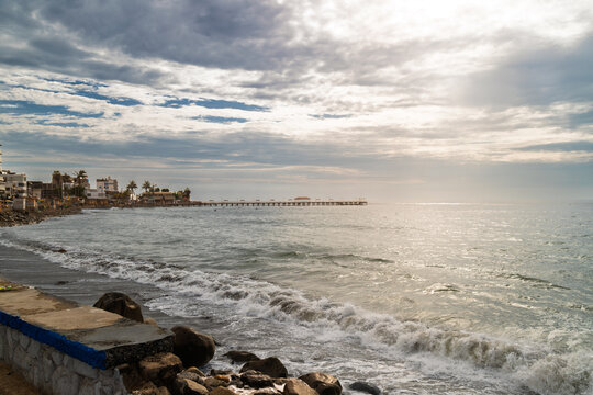 Coastal view of Huanchaco pier with waves crashing on rocky shore and cloudy sky in Peru