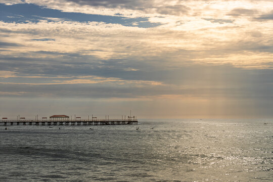 Silhouettes of surfers in the water near Huanchaco pier with golden sun rays and dramatic clouds in Peru