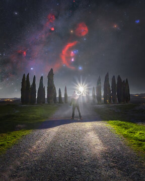 View of a person holding a bright light source in front of the iconic Cipressi di San Quirico d'Orcia cypress trees under a starry night sky in San Quirico d'Orcia, Tuscany, Italy.