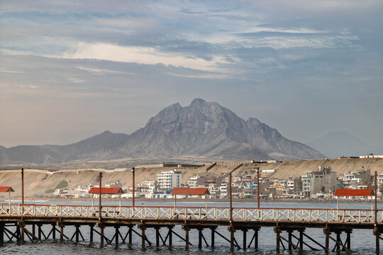 Wooden pier extending over Pacific Ocean with dramatic mountain peak and coastal town of Huanchaco in background