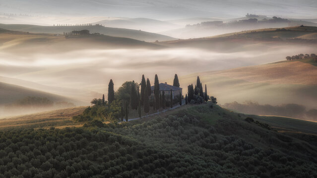 View of Podere Belvedere surrounded by cypress trees on rolling hills covered in morning mist in San Quirico d'Orcia, Toscana, Italy.