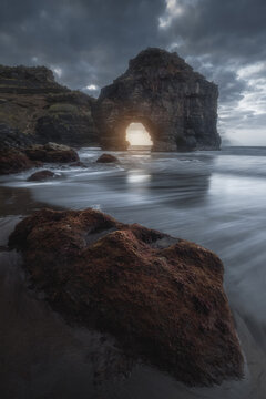View of the Roque Chico volcanic rock formation with a natural arch on the beach, surrounded by dark sea waves and dramatic cloudy sky in Santa Cruz de Tenerife, Canarias, Spain.