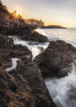 View of the coastal village of Tellaro at sunset with waves crashing against dark rocks and golden light illuminating the colorful houses in Tellaro, Liguria, Italy.