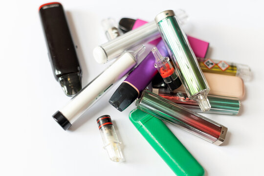 Heap of colorful disposable vapes and pod systems with chemical warning labels, highlighting nicotine addiction, lung damage risks, and health hazards of vaping.