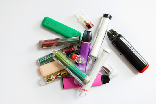 A pile of colorful disposable e-cigarettes and vape devices with toxic warning symbols, representing nicotine addiction, chemical hazards, and health risks of vaping.