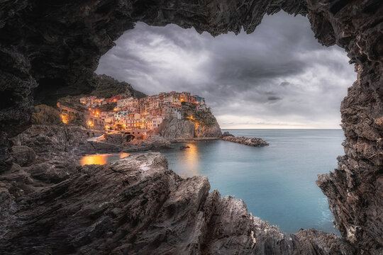 View of the colorful houses of Manarola perched on a rocky cliff overlooking the Ligurian Sea at dusk, framed by a natural rock cave opening in Manarola, Liguria, Italy.