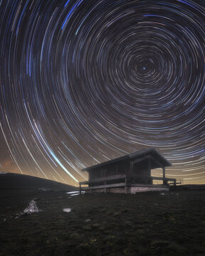 View of Rifugio Belvedere at night with vibrant star trails circling the sky above the wooden cabin in the mountains of Arquata del Tronto, Marche, Italy.