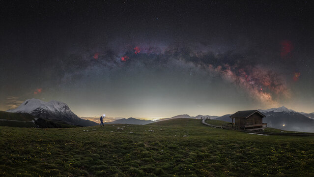View of Rifugio Belvedere under the Milky Way galaxy with a person holding a light in a grassy field with mountains in the background in Arquata del Tronto, Marche, Italy.