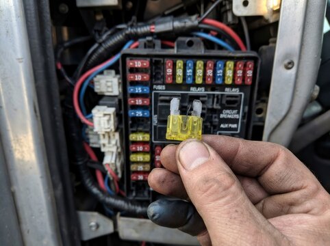 A close-up view of a hand holding a blown automotive fuse with a broken metal element, positioned in front of a vehicle fuse box with various colored electrical fuses and wiring.