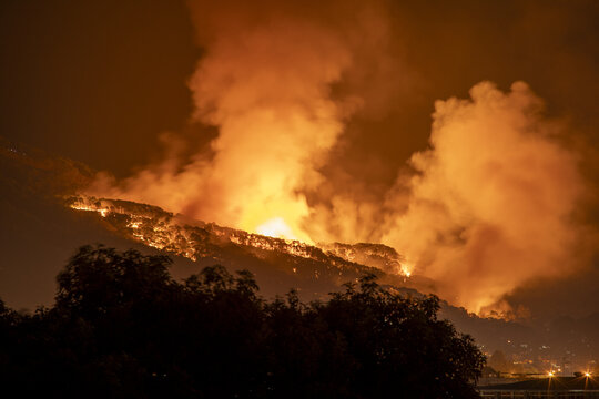 View of a large wildfire burning on a hillside at night, with bright orange flames and thick smoke rising into the dark sky above Uruapan, Michoacan, Mexico.