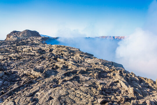 View of the volcanic crater of Erta Ale with jagged solidified lava rock and rising volcanic steam under a clear blue sky in Ethiopia.